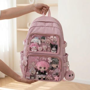 May include: A light pink backpack featuring two clear front display pockets filled with various anime-style plush toys, character pins, and decorative bows. The bag includes side pockets, a top handle, and a fluffy pink pom-pom charm attached to the side.