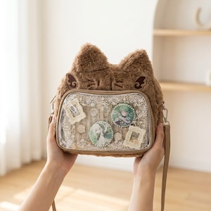 May include: A brown faux fur crossbody bag shaped like a cat head with pointed ears. The front features a clear vinyl window displaying white lace fabric, two circular anime-style character pins, and two small decorative cream-colored frames.