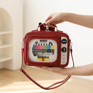 以下が含まれることがあります： ヴィンテージテレビの形をした赤いハンドバッグ。前面には透明な窓があり、NO SIGNALという文字が入ったカラフルなテストパターンが表示されています。側面には2つの黒い回転ノブと3つの小さな銀色のハート型のボタンが付いています。