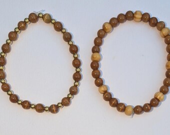 May include: Two circular beaded bracelets. The left bracelet features alternating brown and small metallic gold beads. The right bracelet consists of brown beads interspersed with larger, lighter tan wooden beads in a repeating pattern.