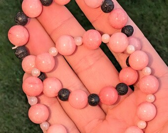 May include: A beaded bracelet featuring alternating smooth, round, light pink beads and textured, porous, black lava beads, accented with smaller, translucent, pale pink spacer beads arranged in a circular pattern on an elastic cord.