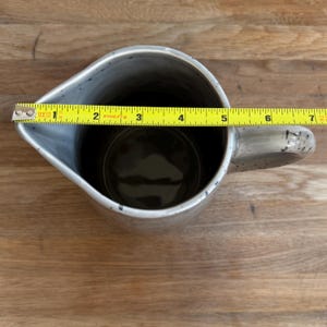 May include: A speckled gray ceramic pitcher with a handle. The pitcher is approximately 6 inches wide, measured with a yellow tape measure. The interior is dark. The pitcher is on a wooden surface.