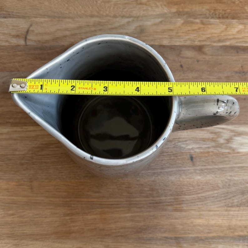 May include: A speckled gray ceramic pitcher with a handle. The pitcher is approximately 6 inches wide, measured with a yellow tape measure. The interior is dark. The pitcher is on a wooden surface.