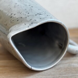 May include: A ceramic pitcher with a light gray speckled glaze. The pitcher has a wide, angled spout and a small handle. The interior is a darker gray. The pitcher is likely used for serving beverages.