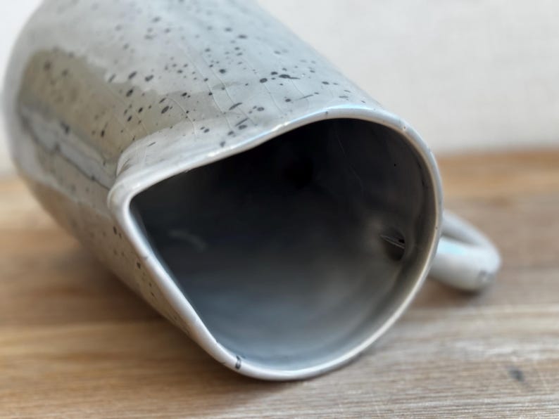 May include: A ceramic pitcher with a light gray speckled glaze. The pitcher has a wide, angled spout and a small handle. The interior is a darker gray. The pitcher is likely used for serving beverages.