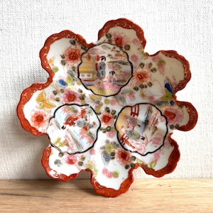 May include: A scalloped, flower-shaped ceramic dish featuring a white base with a vibrant orange border. The surface is decorated with intricate hand-painted scenes of figures in traditional attire, surrounded by colorful floral motifs and butterflies.