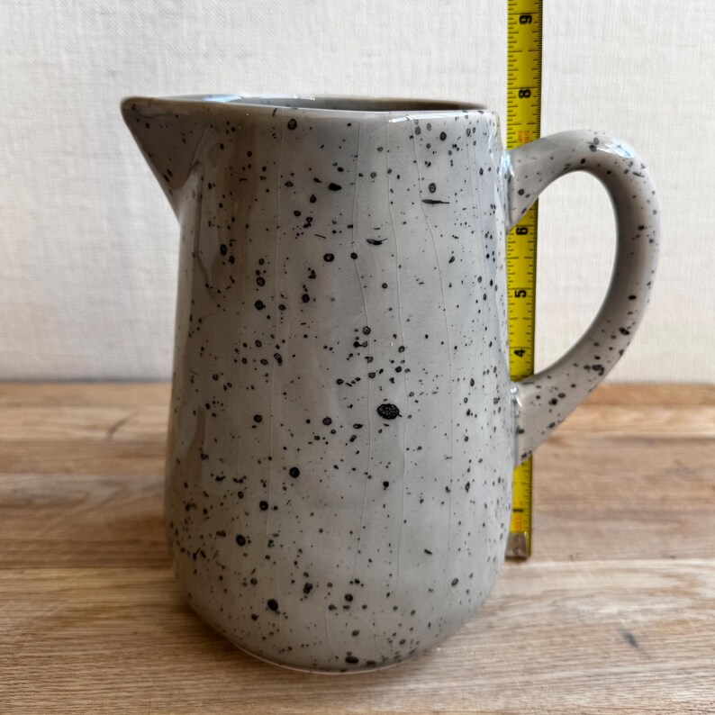 May include: A speckled gray ceramic pitcher with a handle and spout. The pitcher is approximately 8 inches tall. The surface is covered in small black speckles. The pitcher is on a wooden surface.