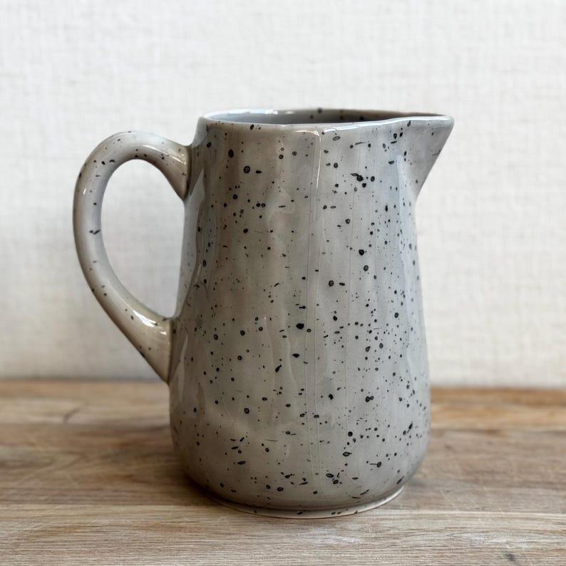 May include: A speckled gray ceramic pitcher with a curved handle and a pouring spout. The pitcher has a light gray base color with small black speckles throughout. The pitcher is sitting on a wooden surface.