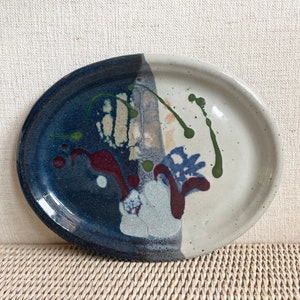 May include: An oval ceramic plate featuring a split color design with a deep navy blue section on the left and a speckled off-white section on the right. The surface is decorated with abstract splatters of dark red and olive green glaze.