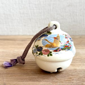 May include: A cream-colored ceramic bell featuring a hand-painted design of a golden crane in flight over blue wave patterns, accented with colorful floral motifs. A thick, braided mauve cord is attached to the top loop of the bell.