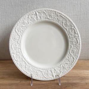 May include: A white ceramic plate with a detailed embossed floral and geometric pattern around the rim. The plate is displayed on a clear stand, showcasing its intricate design. This decorative plate is suitable for serving food.