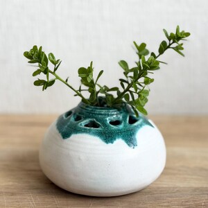 May include: A white ceramic vase with a rounded shape and a teal green rim. The rim features decorative cut-out holes. Green leafy sprigs are arranged inside the vase, creating a natural, decorative display. The vase sits on a wooden surface.