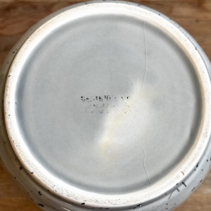 May include: A light gray ceramic teapot with a speckled design. The bottom of the teapot is visible, showing a crack and faded text. The teapot has a handle and a spout.