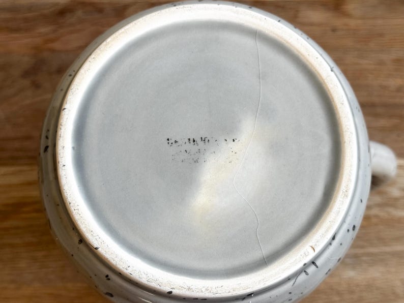 May include: A light gray ceramic teapot with a speckled design. The bottom of the teapot is visible, showing a crack and faded text. The teapot has a handle and a spout.