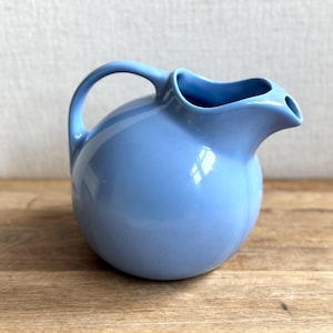 May include: A light blue ceramic pitcher with a rounded body and a curved handle. The pitcher has a spout for pouring and is sitting on a wooden surface. The pitcher is a solid color and has a glossy finish.