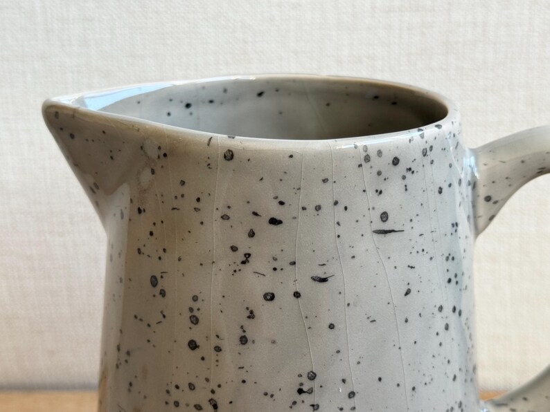 May include: A speckled ceramic pitcher with a spout and handle. The pitcher is a light gray color with small black speckles throughout. The handle is curved and the spout is angled for easy pouring. A handmade kitchenware item.