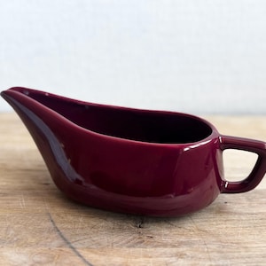 May include: A burgundy-colored gravy boat with a handle. The boat has a long, oval shape with a spout on one end. The handle is a simple loop design. The gravy boat is made of ceramic and has a glossy finish.