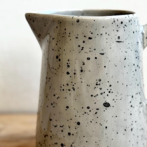 May include: A speckled ceramic pitcher with a spout and handle. The pitcher is off-white with black speckles and visible crack lines. The pitcher is on a wooden surface.