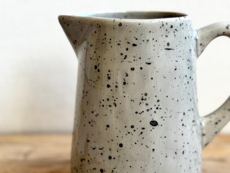 May include: A speckled ceramic pitcher with a spout and handle. The pitcher is off-white with black speckles and visible crack lines. The pitcher is on a wooden surface.