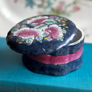 May include: A small, round, navy blue ceramic trinket box with a lid. The lid is decorated with pink and white floral designs and yellow accents. A pink band with a decorative pattern encircles the box.