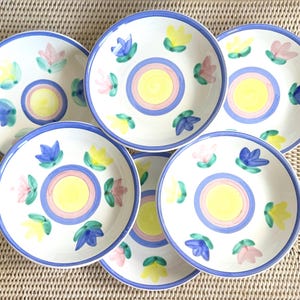 May include: A set of five round ceramic plates featuring a hand-painted design. Each plate has a central yellow circle surrounded by a pink ring and a blue border. The rim is decorated with alternating blue, yellow, and pink floral motifs.