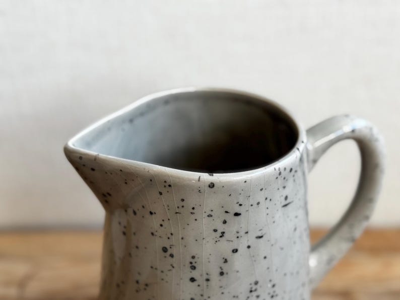 May include: A speckled, off-white ceramic pitcher with a curved handle and a pouring spout. The pitcher has a textured surface with dark gray speckles. The interior is dark. The pitcher is likely used for serving beverages.