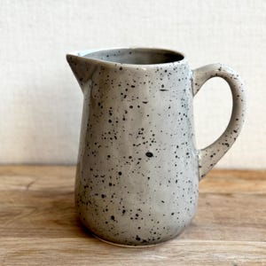 May include: A speckled gray ceramic pitcher with a curved handle and a pouring spout. The pitcher has a light gray base color with small black speckles throughout. The pitcher is sitting on a wooden surface.