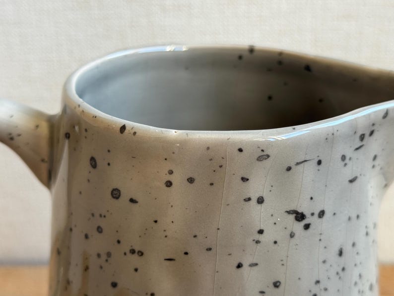 May include: Close-up of a speckled ceramic pitcher with a handle and pouring spout. The pitcher is a light gray color with small, dark brown speckles. The interior is a lighter gray.