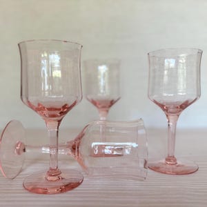 May include: Four pink-tinted stemmed glasses. Three glasses are upright, and one is lying on its side. The glasses have a textured, slightly irregular rim and a rounded base. The stems and bases are a deeper shade of pink.