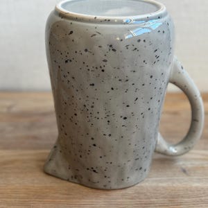 May include: A speckled gray ceramic mug with a unique, asymmetrical shape. The mug features a rounded handle and a speckled pattern of black dots across its surface. The interior is a lighter gray.