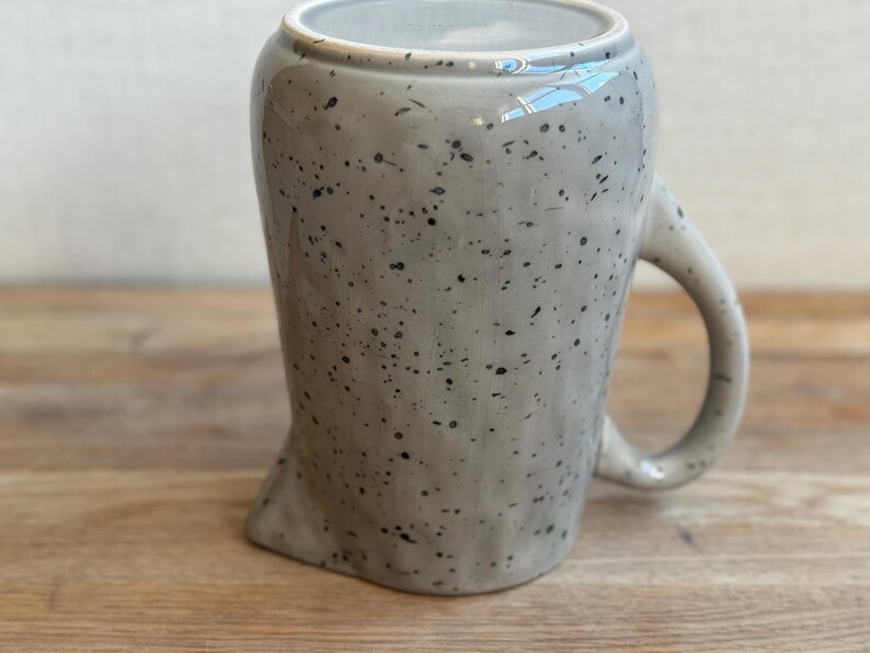 May include: A speckled gray ceramic mug with a unique, asymmetrical shape. The mug features a rounded handle and a speckled pattern of black dots across its surface. The interior is a lighter gray.