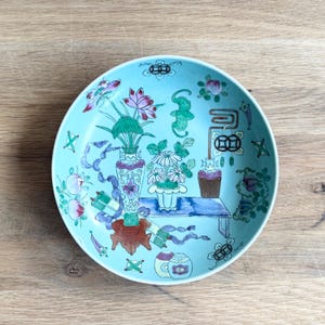 May include: A round ceramic plate with a light blue background featuring a hand-painted design of a vase with pink lotus flowers, a potted plant with white daisies, a small table, and various decorative symbols including coins and scrolls.
