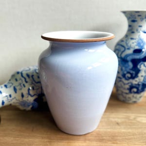 May include: A light blue ceramic vase with a white interior and a brown rim. The vase has a rounded body and a wide opening. Other decorative vases are in the background.