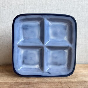 May include: A square, divided serving dish with four compartments. The dish is a gradient of blue hues, with a darker blue rim. The compartments are square and the dish sits on a wooden surface.