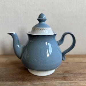 May include: A blue ceramic teapot with a matching lid and handle. The teapot has a white base and a decorative band around the lid. The spout and handle curve gracefully. The teapot is sitting on a wooden surface.