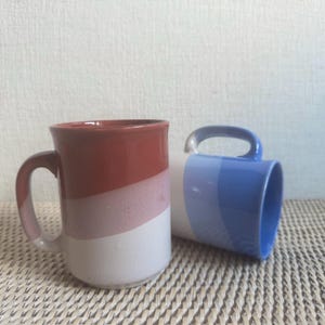 May include: Two ceramic mugs with a diagonal color block design. One mug features a gradient of red, pink, and white. The other mug has a gradient of blue and white. Both mugs have a curved handle.