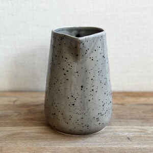 May include: A gray ceramic pitcher with a speckled pattern. The pitcher has a tapered shape and a spout for pouring. The surface is covered in small black dots, giving it a rustic appearance. The pitcher is sitting on a wooden surface.