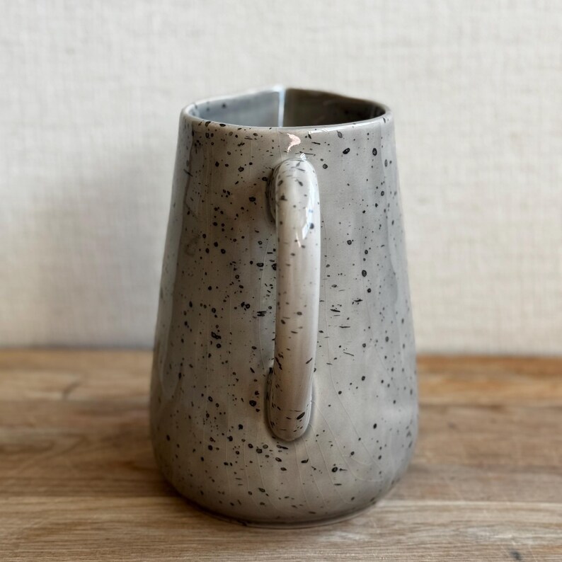 May include: A gray ceramic pitcher with a speckled design. The pitcher has a handle and a slightly flared shape. The interior is a lighter gray. The pitcher is sitting on a wooden surface.