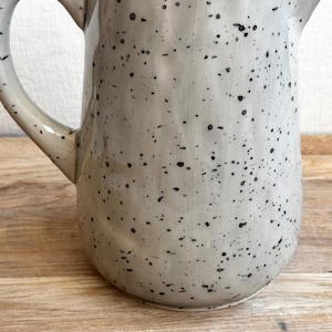 May include: A speckled ceramic pitcher with a handle and spout. The pitcher is light gray with black speckles, and sits on a wooden surface. The handle curves gracefully from the top to the side of the pitcher.