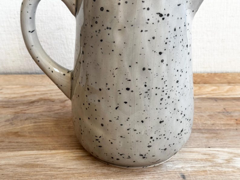 May include: A speckled ceramic pitcher with a handle and spout. The pitcher is light gray with black speckles, and sits on a wooden surface. The handle curves gracefully from the top to the side of the pitcher.