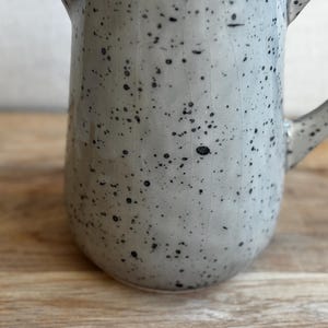 May include: A speckled ceramic pitcher in a light gray color with black speckles. The pitcher has a handle and a spout. The background is a wooden surface.