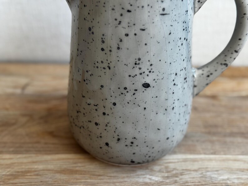 May include: A speckled ceramic pitcher in a light gray color with black speckles. The pitcher has a handle and a spout. The background is a wooden surface.