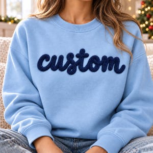 May include: A light blue crewneck sweatshirt featuring the word custom written in dark blue, textured, raised chenille-style lettering across the chest. The garment has long sleeves with ribbed cuffs and a ribbed neckline.