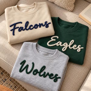 May include: Three crewneck sweatshirts featuring textured, raised lettering. A beige sweatshirt with dark blue 'Falcons' text, a dark green sweatshirt with white 'Eagles' text, and a heather gray sweatshirt with dark green 'Wolves' text.
