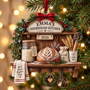 May include: A wooden Christmas ornament shaped like a kitchen shelf featuring a sign reading EMMA'S SOURDOUGH KITCHEN and 2026. It includes a sourdough starter jar, a loaf of bread, a flour sack, wheat stalks, a whisk, and a towel labeled Good Bread Brighter Days.