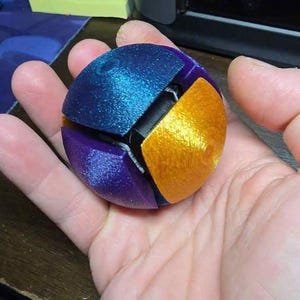 May include: A spherical, multi-coloured fidget toy featuring interlocking segments in metallic blue, purple, and gold. The object has a textured, 3D-printed finish with visible layer lines, and the segments are arranged to form a round shape.