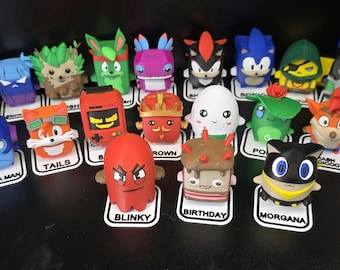 May include: A collection of small, colourful, blocky character figurines arranged on a dark surface. Each figure stands on a white base with a printed name tag, including characters like Mega Man, Tails, Blinky, Birthday, and Morgana, alongside a grey chest.
