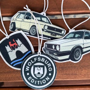May include: Four hanging air fresheners featuring two white Volkswagen Golf car illustrations, a black shield emblem with a red wolf and castle graphic, and a circular black badge with the text Wolfsburg Edition and a castle icon.