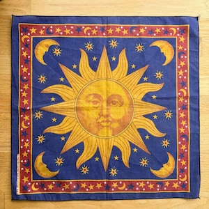 May include: A square blue bandana featuring a large central yellow sun with a human face, surrounded by yellow stars and crescent moons. The border is a red band decorated with a repeating pattern of small yellow stars and blue crescent moons.