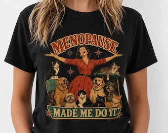 Puede incluir: Una camiseta negra de manga corta con un estampado gráfico de una persona con vestido rojo rodeada de varios gatos y perros. El texto "MENOPAUSE" aparece arqueado arriba y "MADE ME DO IT" está impreso en una cinta en la parte inferior.
